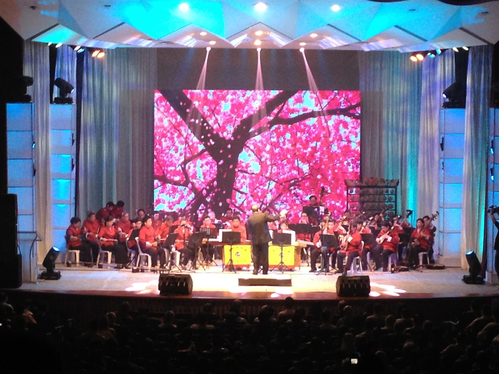 Orchestra performing on a large stage with a central LED screen and blue lighting